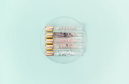 5 Crystal Roller Bottles with Essential Oil Blends and gold caps lined up on a clear glass plate with a pale aqua backdrop