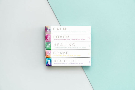 5 boxes with watercolor ends that hold Crystal Essential Oil Blends in Roller Bottles on a backdrop that has half white and half aqua on a diag.