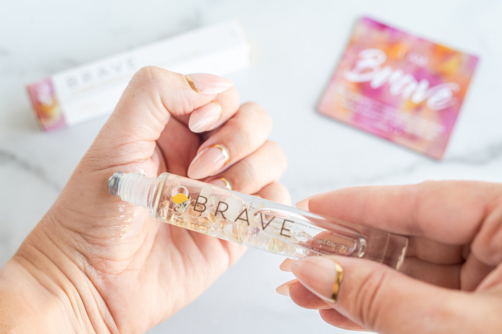 Hand holding a small bottle labeled 'Brave' with colorful packaging in the background.