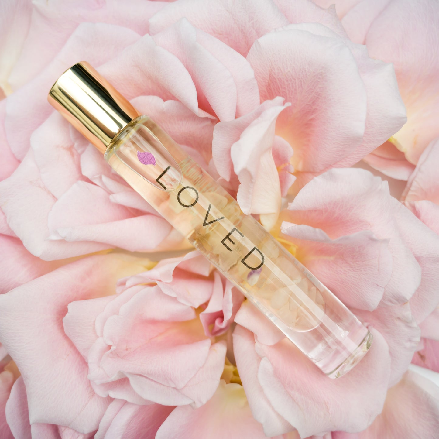 A Loved Crystal Roller Bottle Blend in glass bottle with gold cap containing rose quartz crystals laying on pink roses