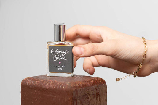 Hand holding a perfume bottle labeled 'Bunny Kisses' on a wooden surface with a blurred natural background