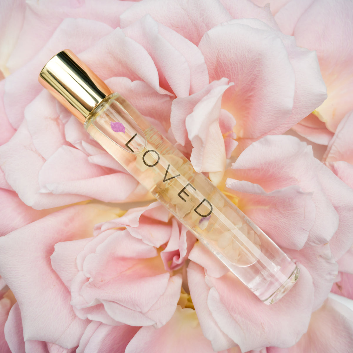 A glass roller bottle with gold cap labeled LOVED on a light pink rose with the petals spread apart.