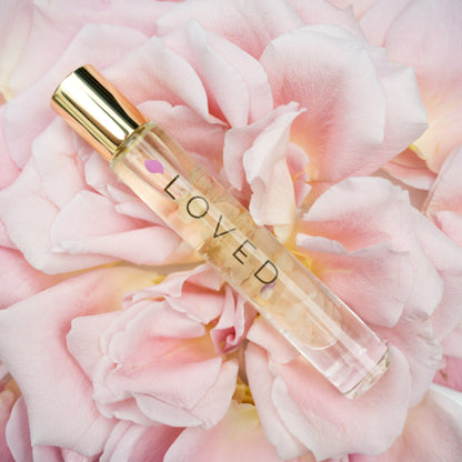 A glass roller bottle with gold cap labeled LOVED on a light pink rose with the petals spread apart.