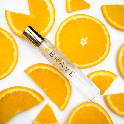 A glass roller bottle with gold cap labeled BRAVE and orange slices in the background