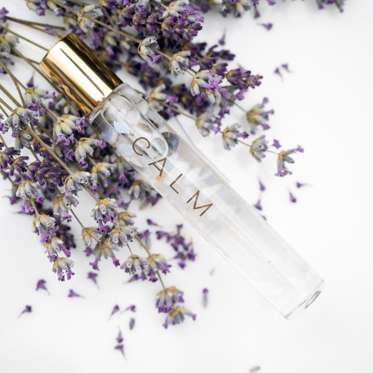 A glass roller bottle with gold cap labeled CALM with a sprig of lavender and lavender buds in the background