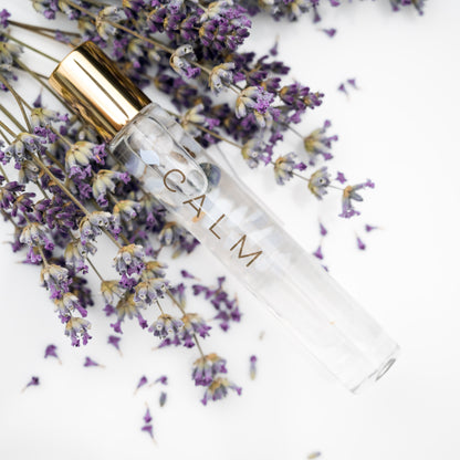 A glass roller bottle with gold cap labeled CALM with a sprig of lavender and lavender buds in the background