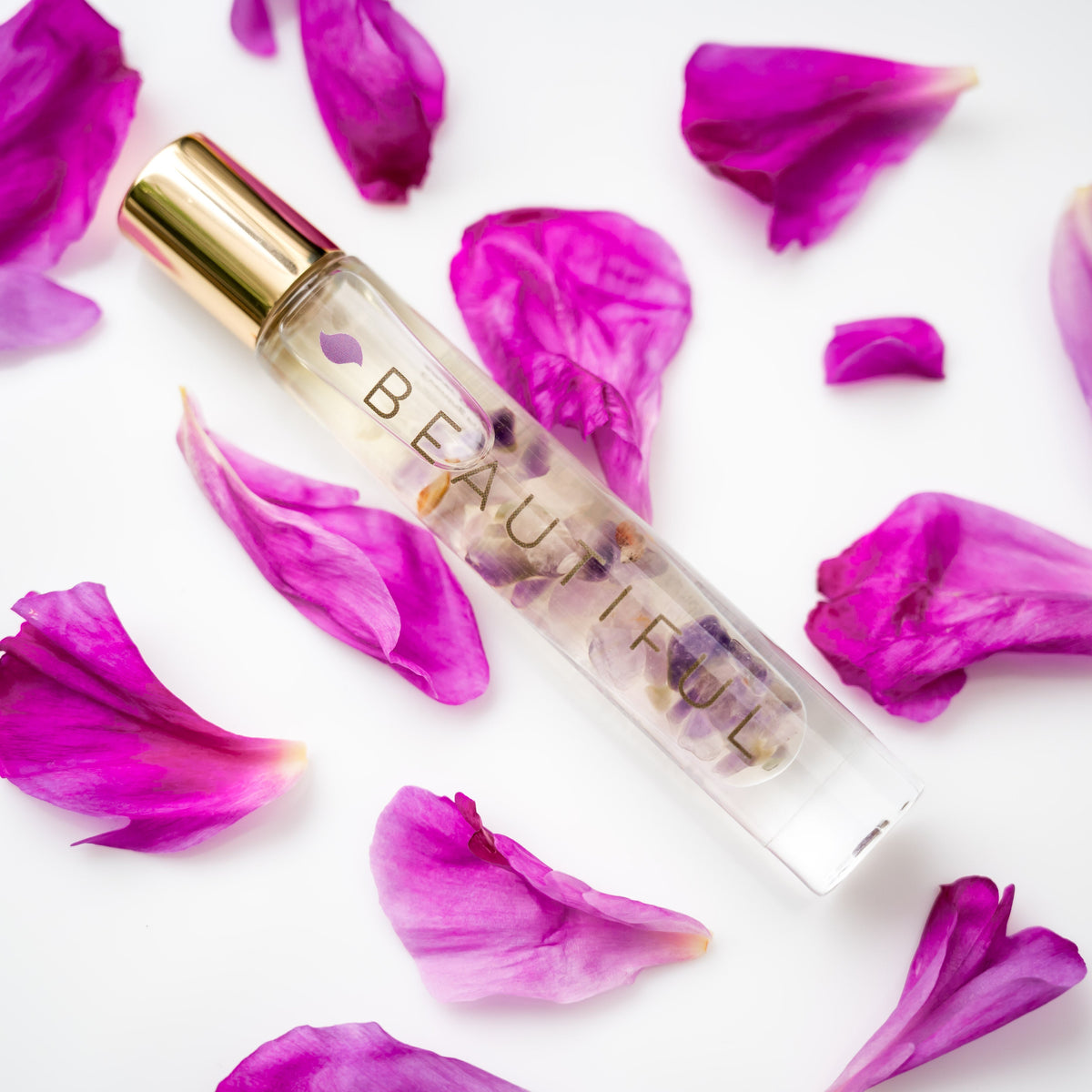A glass roller bottle with gold cap labeled BEAUTIFUL with purple flower petals on the background