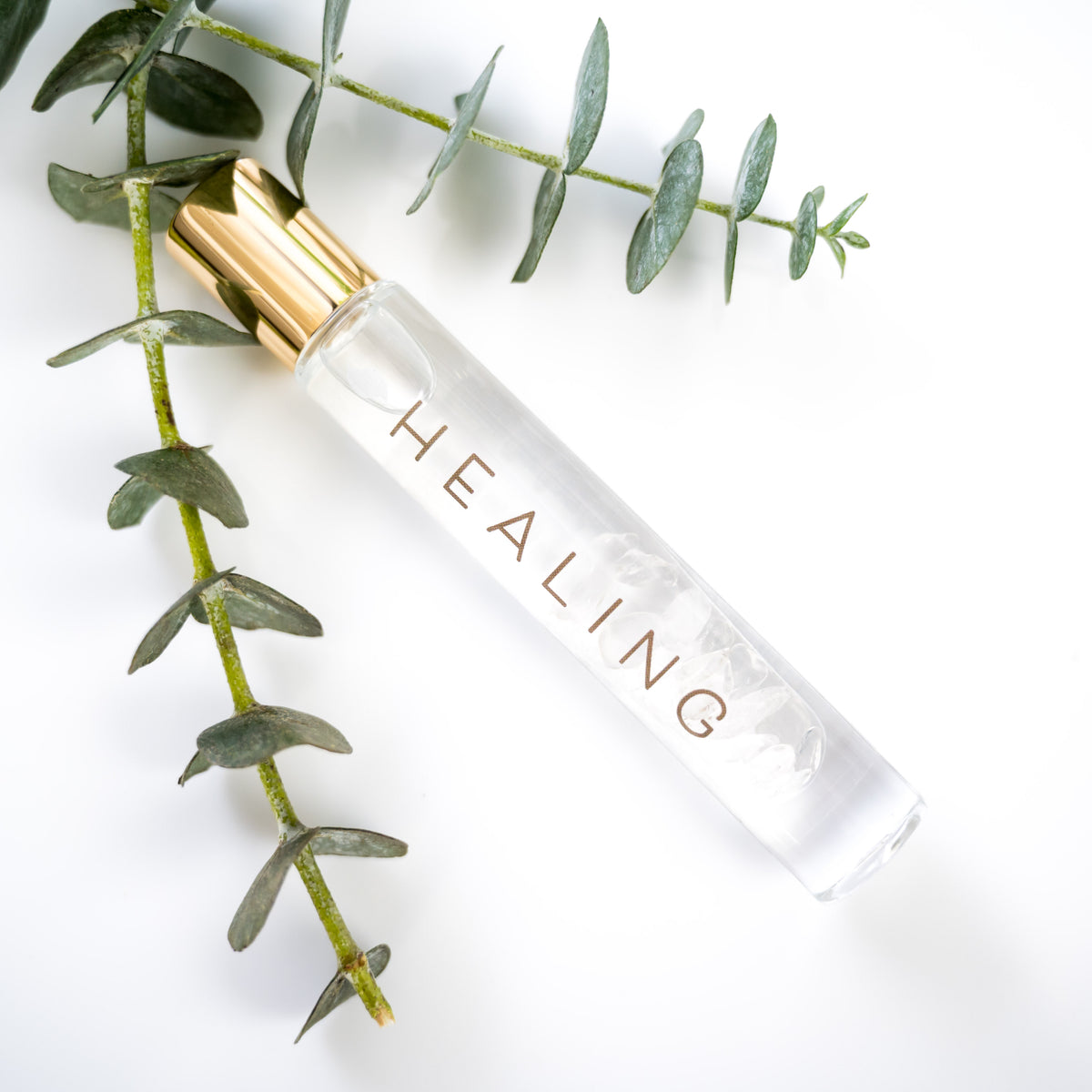 A glass roller bottle with gold cap labeled HEALING with a sprig of eucalyptus on a white background