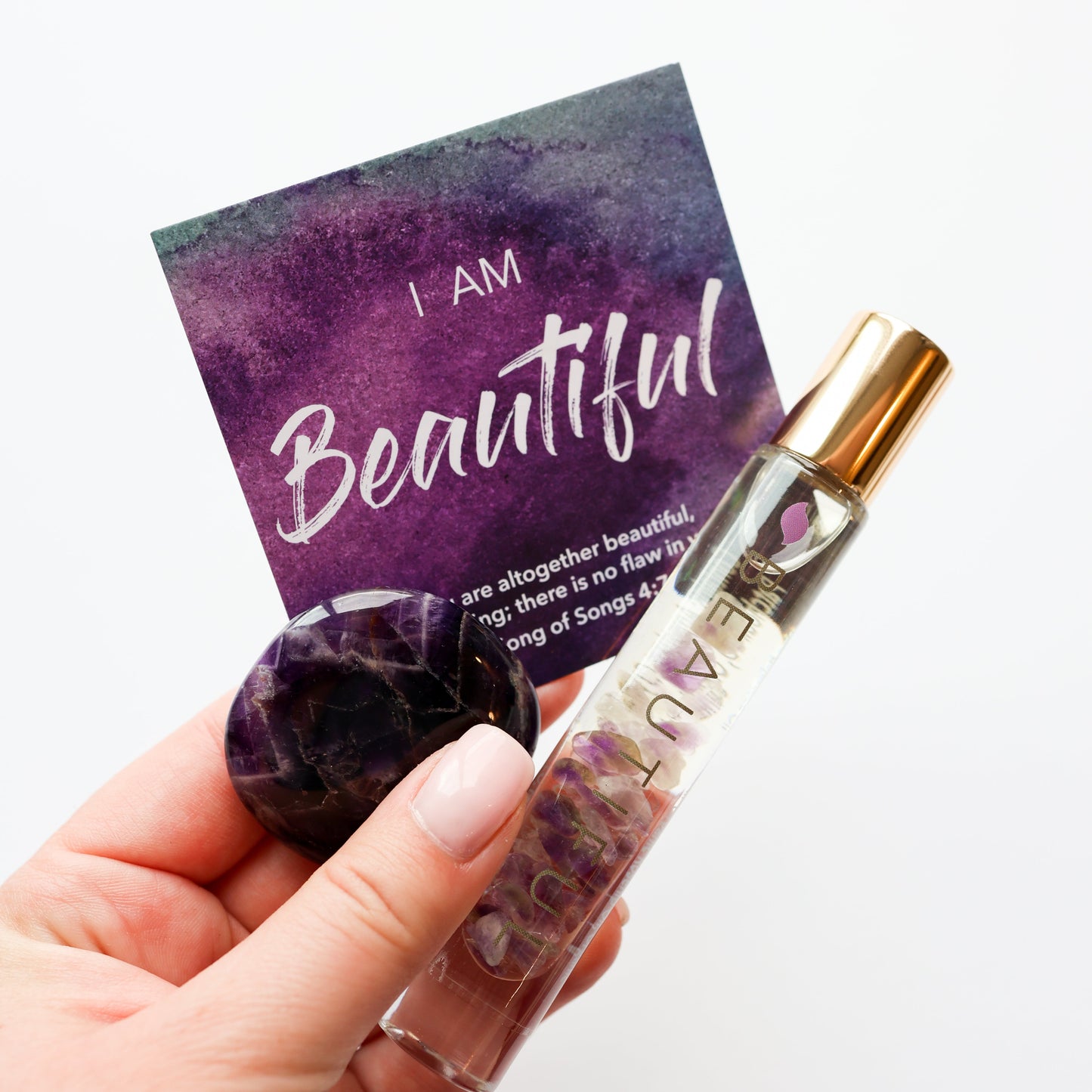 Hand holding a small bottle with a purple amethyst crystal, a card with 'I AM Beautiful' text, on a white background.