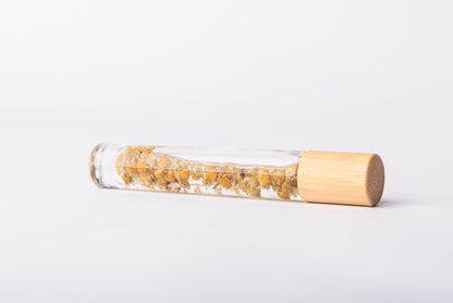 A clear glass roller bottle with Bamboo lid laying on it's side filled with organic chamomile flower buds